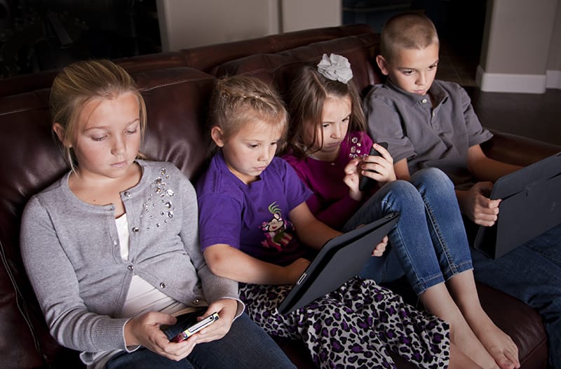 Children sitting on the couch, each looking at a mobile phone or tablet